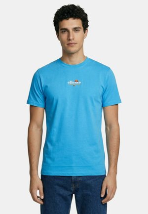 Homme portant un t-shirt bleu vif à manches courtes avec le logo "ellesse", associé à un jean bleu, debout devant un fond blanc uni.
