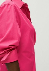 Lyserød skjorte med krave og puffede ærmer, der har en glat tekstur og en struktureret silhuet, vist fra siden.