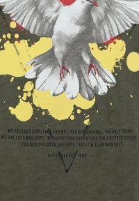 Camisa de color verde oliva con un estampado de pájaros grises y acentos rojos. Fondos salpicados en amarillo y negro. Texto en negrita en fuente negra en la parte inferior.