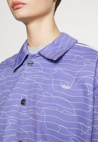 Lila knäppt jacka med ett vågigt vitt linjemönster och en liten logotyp. Har en krage och svarta knappstängningar; tyget ser slätt ut.