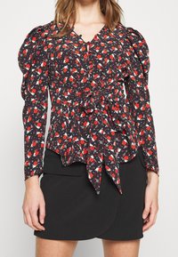 Blouse noire à motifs floraux rouges et blancs, manches bouffantes, taille cintrée et détail noué à l'avant. Associée à une jupe noire.