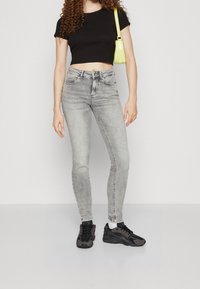 Camiseta negra recortada, jeans ajustados gris claro y zapatillas negras. La modelo sostiene un pequeño bolso verde lima. Diseño simple, estilo casual.