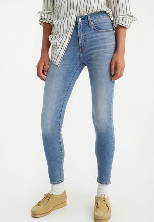 Persoon die lichtblauwe skinny jeans draagt, een wit overhemd met zwarte verticale strepen, witte sokken en beige suède schoenen.