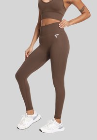 Kobieta w brązowych legginsach z wysokim stanem i dopasowanym staniku sportowym, w białych tenisówkach, stojąca na tle jasnoszarym.