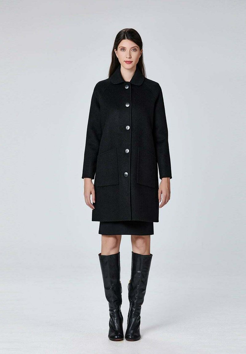 Manteau en laine noir avec un col rond, fermeture par bouton à l'avant, deux poches avant et manches longues. Porté par-dessus une jupe noire et des bottes noires montantes.