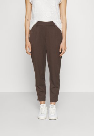 Kvinde iført mørkebrune smalle bukser, hvid blonder top uden ærmer og hvide sneakers, stående foran en ensartet lys baggrund.