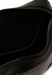 Interior de bolso de cuero negro con una textura suave y cierre de cremallera. Presenta un forro de tela negra, amplio y sin adornos.