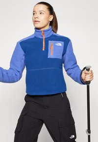 Plavi flis pulover s polu-zip ovratnikom, kontrastnom svijetloplavom džepom na prsima i narančastim detaljima na patentnom zatvaraču. Tekstuirana tkanina za dodatnu toplinu.