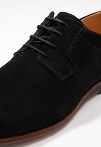 Chaussure en daim noire avec un design traditionnel à lacets, présentant des coutures contrastées et une semelle lisse marron. Vue rapprochée mettant en évidence la texture.