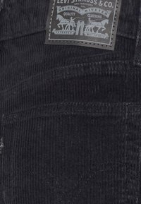 Tissu en velours côtelé noir avec des rainures verticales, présentant une étiquette Levi Strauss & Co. représentant des chevaux et des personnages. Des détails de couture sont visibles.