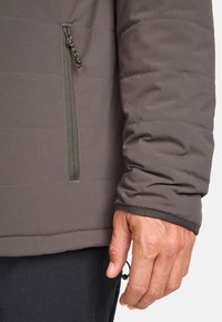 Chaqueta acolchada gris con un bolsillo con cremallera en el brazo izquierdo, que presenta puños elásticos y una textura de tela suave. Ajustador en el dobladillo.