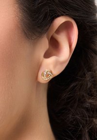 Kolczyki w odcieniu złota w kształcie węzła z plecionym wzorem, zdobione przezroczystymi kamieniami i polerowanym wykończeniem, pewnie przymocowane do płatka ucha.