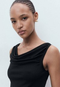 Zwarte mouwloze top met gedrapeerde halslijn. Soepele textuur, aansluitend ontwerp en een lichte glans. Model heeft gevlochten haar en oorbellen met ringen.