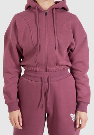 Mujer que lleva un conjunto a juego de sudadera con capucha y pantalones deportivos de color malva con cordones y un pequeño diseño de corazón blanco en el muslo izquierdo.