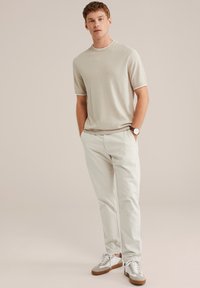 Camicia beige a maniche corte con texture, colletto e polsini bianchi a contrasto, abbinata a pantaloni crema e sneakers chiare con dettagli marroni.