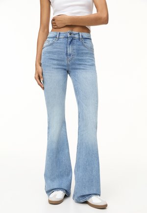 Personne portant un jean évasé taille haute bleu clair, des baskets blanches et un débardeur court blanc, debout devant un fond blanc.