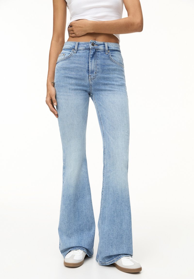 Personne portant un jean évasé taille haute bleu clair, des baskets blanches et un débardeur court blanc, debout devant un fond blanc.
