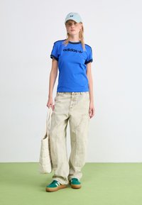 Blå bomull T-skjorte med svart adidas-logo, beige baggy bukser, grønne semskede sneakers og en lyseblå caps med et preg.