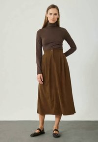 Bruine coltrui en een hoge taille, enkel-lange bruine geplooide rok met een knoopdetail. Model draagt zwarte platte schoenen.