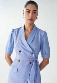 Robe bleu clair avec des rayures verticales blanches, devant croisé à double boutonnage, manches courtes bouffantes, lien sur le côté et deux poches à l'avant. Détails en boutons dorés.