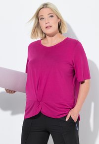 Camiseta corta de manga corta en fucsia con detalle de nudo en la parte frontal, hecha de un material suave. Combinada con pantalones negros con bolsillos con cremallera.