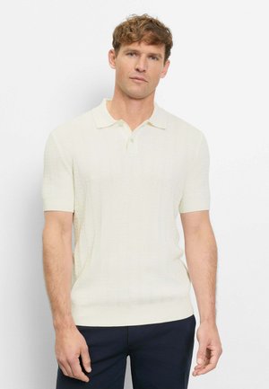 Man with short curly hair wearing a short-sleeve cream knit polo shirt and dark pants, standing against a plain white background.