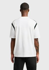 T-shirt bianco a maniche corte con accenti neri sulle spalle, texture morbida e vestibilità rilassata. Tatuaggi visibili sul braccio.