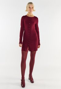 Kobieta stojąca w bordowej sukience z guzikami, pasujących krytych rajstopach i lakierowanych butach Mary Jane na jednolitym białym tle.