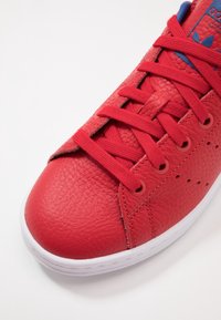 Rode leren sneaker met een gestructureerd oppervlak, afgeronde neus en platte veters. Heeft blauwe accenten en perforaties langs de zijkant. Witte zool.