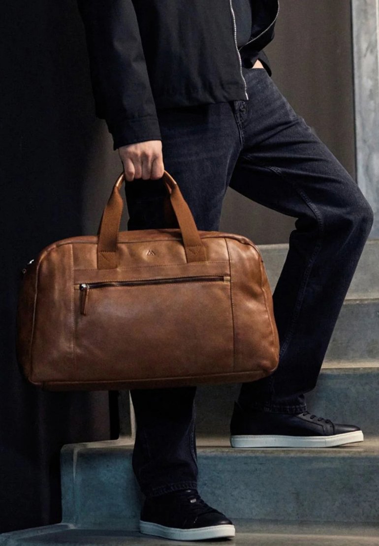 Πρόσωπο κρατάει μία καφέ δερμάτινη τσάντα ενώ στέκεται σε γκρίζα σκαλιά, φορώντας μαύρα τζιν, μαύρο μπουφάν και μαύρα αθλητικά παπούτσια με λευκές σόλες.