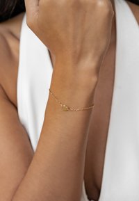 Pulsera de oro con un colgante en forma de corazón sobre una delicada cadena, llevada en una muñeca esbelta, con superficies lisas y acabado brillante.