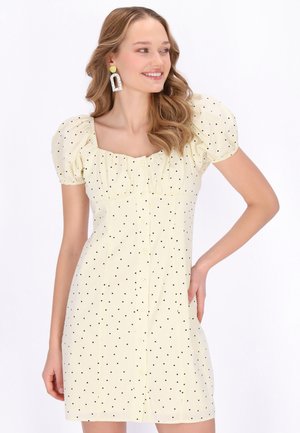 Nainen aaltoilevine hiuksineen, pukeutunut keltaiseen, mustilla pilkuilla koristeltuun lyhythihaiseen mekkoon, jossa on napit edessä, hymyilevä ja katsomassa oikealle, käsi lanteilla.