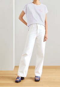 Camiseta blanca a rayas con mangas cortas y un detalle bordado en rosa, combinada con pantalones vaqueros blancos de pierna ancha y sandalias de tacón cuadrado azul con tiras.