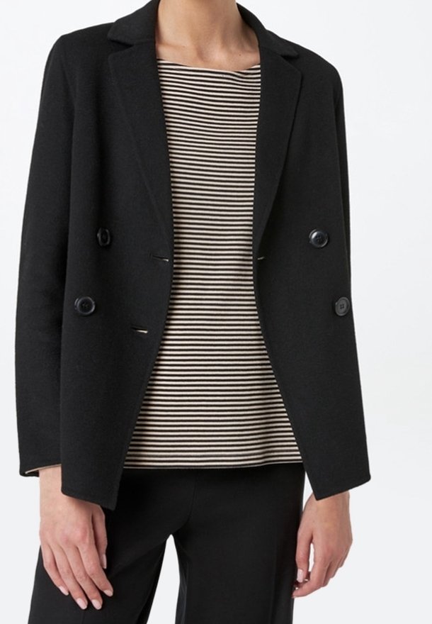 Personne portant un blazer noir croisé sur une chemise rayée noir et blanc et un pantalon noir, debout devant un fond blanc.