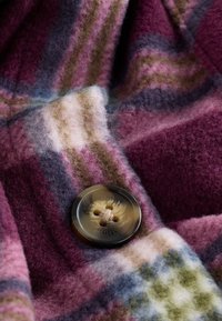 Blødt uldstof med rudemønster i bordeaux, lilla, grønt, hvidt og blåt, prydet med en stor rund brun knap syet med beige tråd.