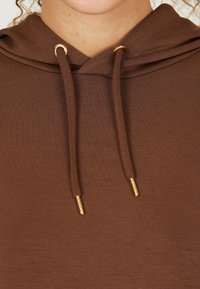Hoodie marron avec une texture douce, doté d'une capuche à cordon et d'œillets dorés. Le tissu semble léger et ajusté.