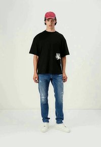 T-shirt preta oversized com um design gráfico branco na frente, combinada com jeans azuis rasgados e ténis brancos. Boné rosa na modelo.