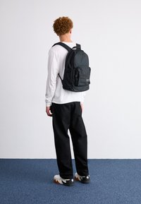 Czarny plecak z tkaniny z przednią kieszenią, regulowanymi ramiączkami i uchwytem na górze, noszony na białej bluzce z długim rękawem i czarnych spodniach.