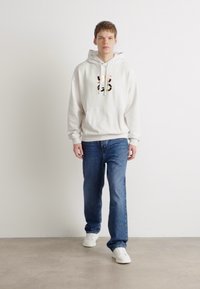 Sudadera blanca con estampado gráfico, corte relajado. Combinada con jeans de mezclilla azules y zapatillas blancas, de pie frente a un fondo liso.