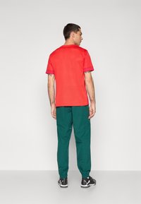 Rød træningsshirt med korte ærmer, parret med løse grønne bukser og sorte sneakers. Enkel design, glat tekstur og ingen fremtrædende mønstre.