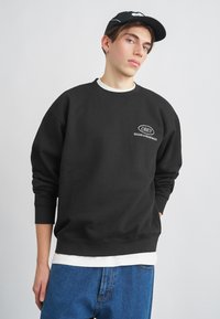 Črna majica s kratkimi rokavi s krožnim izrezom in rebrastimi manšetami, na kateri je bel logotip "OBEY". Oblačena je čez belo večplastno srajco z modrimi kavbojkami.