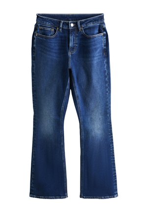 Kvinde stående barfodet med den ene hånd bag hovedet, iført en brun kortærmet top og mørkeblå straight-leg jeans.