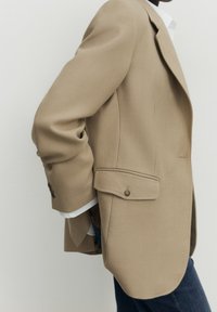 Blazer beige con un design strutturato, revers a punta, due bottoni sui polsini e tasche laterali. Abbinato a una camicia bianca e jeans scuri.