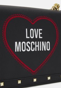Svart handväska med "LOVE MOSCHINO" i vit broderi, omgiven av en röd hjärtstickning. Guldhårdvara och nitar längs botten.