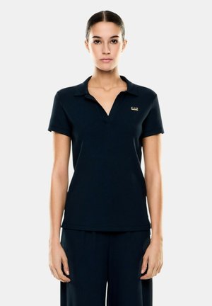 Donna con capelli scuri raccolti, indossa una polo nera a maniche corte e pantaloni abbinati, in piedi davanti a uno sfondo chiaro uniforme.