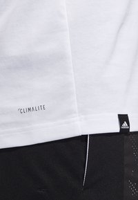Camiseta de algodón blanca con etiqueta 'CLIMALITE' en la costura lateral y etiqueta de logo negra de Adidas en el dobladillo inferior. Diseño sencillo, textura suave.