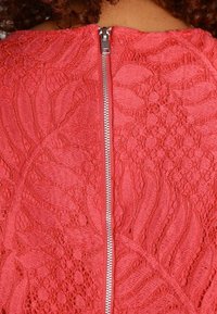 Widok z tyłu osoby w dopasowanej czerwonej koronkowej sukience z widocznym srebrnym zamkiem błyskawicznym biegnącym wzdłuż środka pleców.