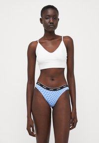 White bralette with thin straps, paired with blue thong featuring a repeating white "HUGO" logo on the waistband and smooth texture.