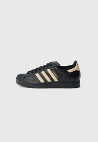 Černá teniska Adidas Superstar s béžovými pruhy, designem špičky s vnější ochranou a značkovým poutkem na patě na bílém pozadí, boční pohled.