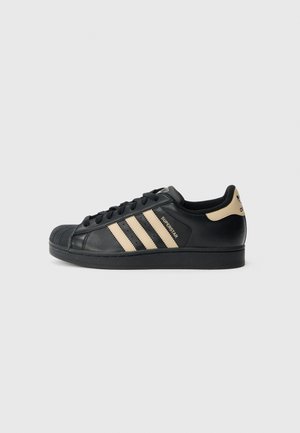 Schwarzer Adidas Superstar Sneaker mit beigen Streifen, Shell-Toe-Design und gebrandetem Fersenlasche auf weißem Hintergrund, Seitenansicht.